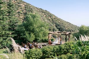 Outdoor wedding ceremony in a lush green valley with mountain backdrop and wooden pergola altar.