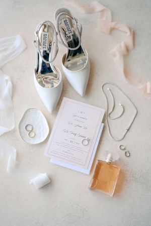Elegant wedding flat lay with white heels, invitation, perfume, rings, and pearl necklace on neutral background.