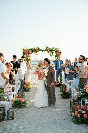 A beach wedding ceremony with guests gathered around a floral arch and white chairs during sunset.