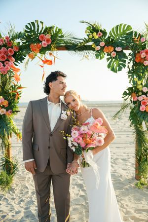 Tropical beach wedding ceremony setup with floral arch featuring monstera leaves and coral blooms.