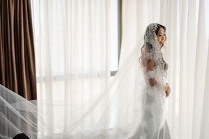Elegant bride in lace wedding dress and long veil standing by sheer curtained window in soft natural light.