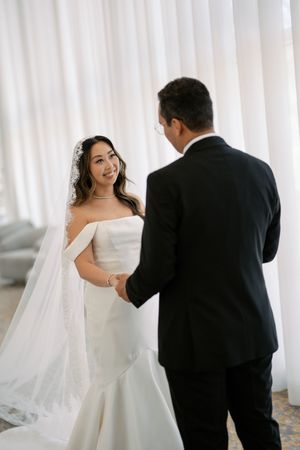 Bride in off-shoulder white gown and veil holds hands with groom in black tuxedo during romantic first look.