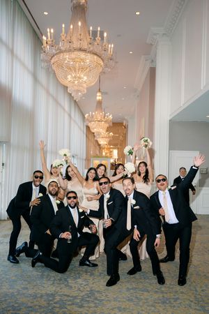Wedding party celebrating in elegant ballroom with crystal chandeliers, groomsmen in black suits and bridesmaids in champagne gowns.