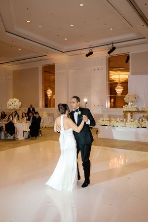 Bride and groom share their first dance on a white marble floor in an elegant ballroom with chandelier lighting.