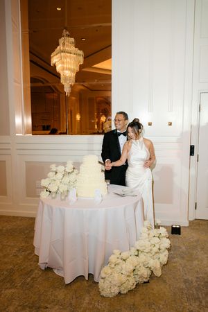 Bride and groom cutting white wedding cake adorned with roses in an elegant ballroom with chandelier.