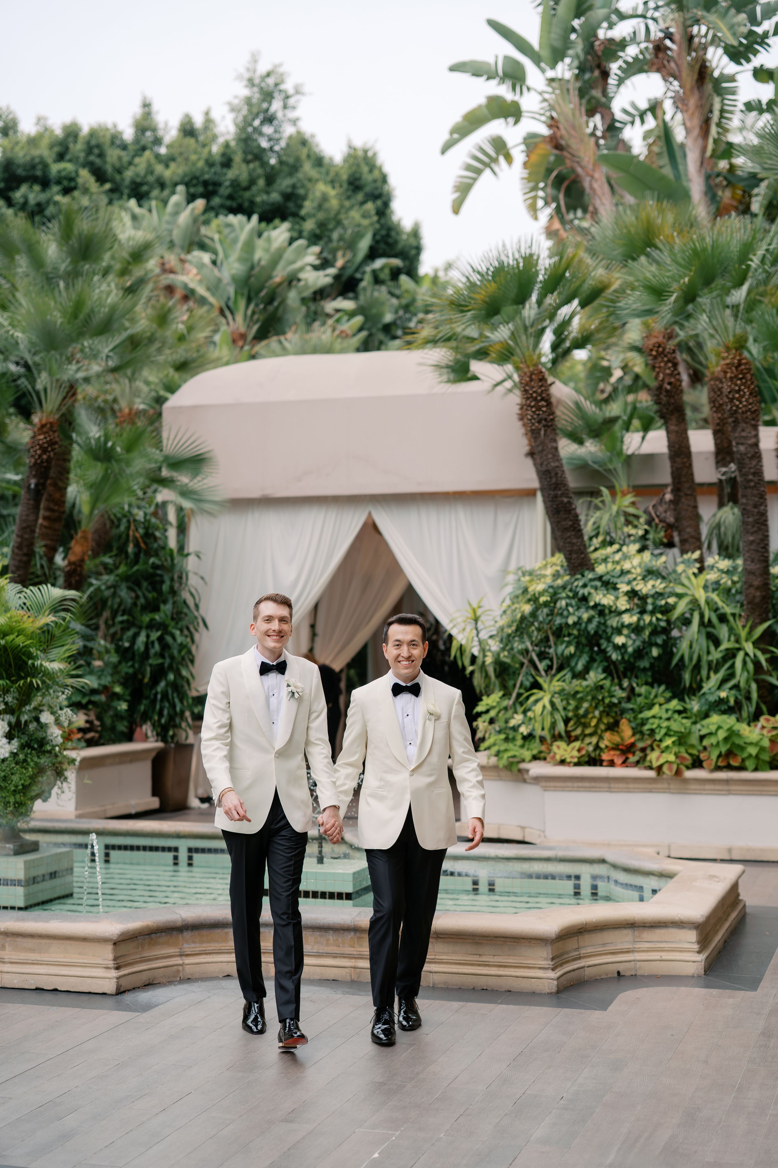 Two grooms in white tuxedo jackets and black bow ties hold hands outdoors near a fountain surrounded by palm trees.