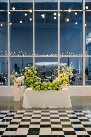 Elegant wedding reception setup with string lights and floral arrangements against large city view windows.