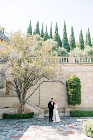 A couple stands in formal attire at a Mediterranean-style courtyard with tall cypress trees and stone walls in the background.
