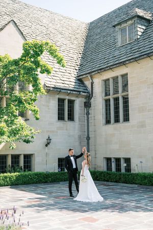 A couple in wedding attire stands together in the courtyard of an elegant stone building with slate roof and climbing vines.