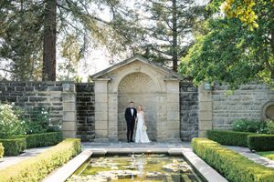 Newlyweds share a romantic moment in a garden walkway surrounded by lush greenery and stone architecture.