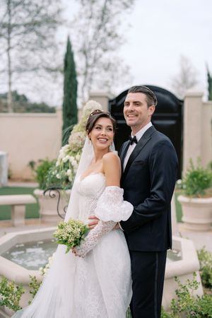Bride in strapless lace gown with veil and groom in black tuxedo posing by fountain in elegant outdoor garden.