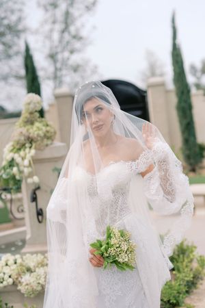 Bride in lace off-shoulder gown and veil holds white bouquet outdoors near elegant stone architecture and cypress trees.