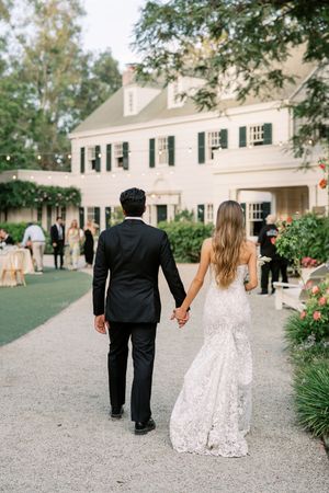 Bride and groom walk hand in hand toward elegant white manor house at outdoor wedding reception.