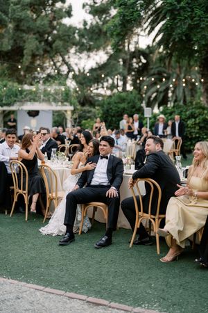 Bride and groom laughing with guests at an elegant outdoor wedding reception with string lights and lush greenery.