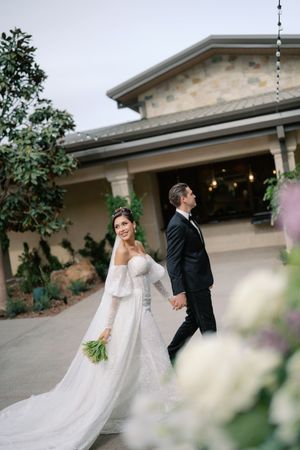 Bride in off-shoulder gown holding bouquet walks hand-in-hand with groom in black tuxedo outside elegant venue.