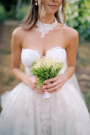 Bride in strapless white lace gown holding small lily of the valley bouquet with green leaves outdoors.