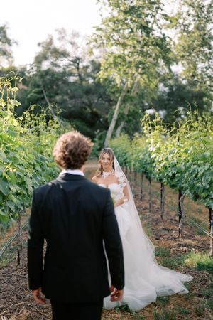 Bride in white gown smiles at groom in black suit walking through lush vineyard rows on their wedding day.