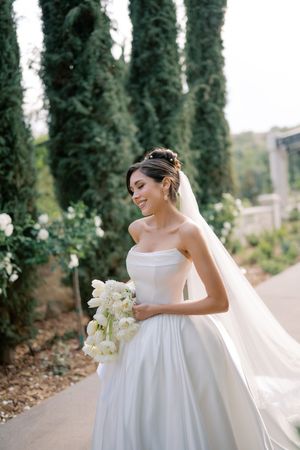 Smiling bride in strapless white ball gown holding white tulip bouquet with veil flowing, posing outdoors near tall cypress trees.