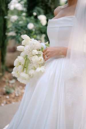 Bride in off-shoulder white gown holding elegant bouquet of white tulips and hydrangeas outdoors.