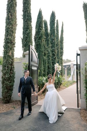 Joyful newlyweds exit through elegant gates lined with tall cypress trees, bride raising bouquet.