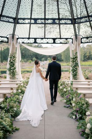 Bride and groom walk hand-in-hand under an elegant gazebo adorned with white draping and floral arrangements at their wedding.
