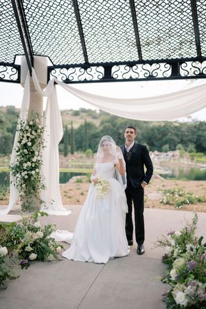 Bride and groom stand under elegant iron pergola with white draping and lush floral arrangements outdoors.