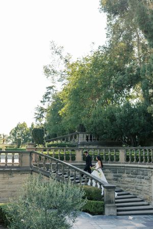 A wedding couple walks up elegant stone steps surrounded by lush greenery and architectural details.
