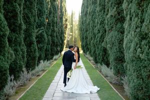 Bride and groom share a romantic kiss along a tree-lined garden path with tall cypress trees at their wedding.