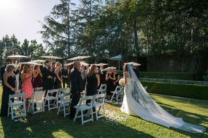Bride in a long white gown and veil walks down the aisle between white chairs toward guests at an outdoor garden wedding ceremony.
