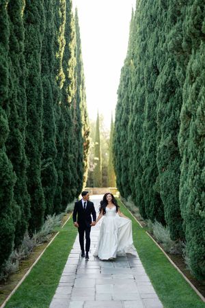 Wedding couple strolling down a dramatic cypress tree-lined pathway in formal attire.