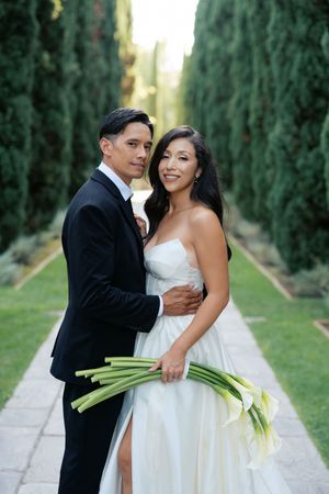 A couple in wedding attire share romantic moments while walking along a cypress tree-lined path.