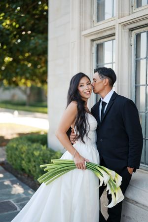 Formal wedding couple standing close together against a white building exterior with classic windows.