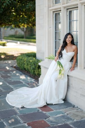 A person in a flowing white wedding dress poses elegantly against a light-colored stone wall on a patio.