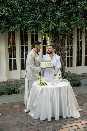 Two people in light colored suits cutting a wedding cake at an outdoor reception.