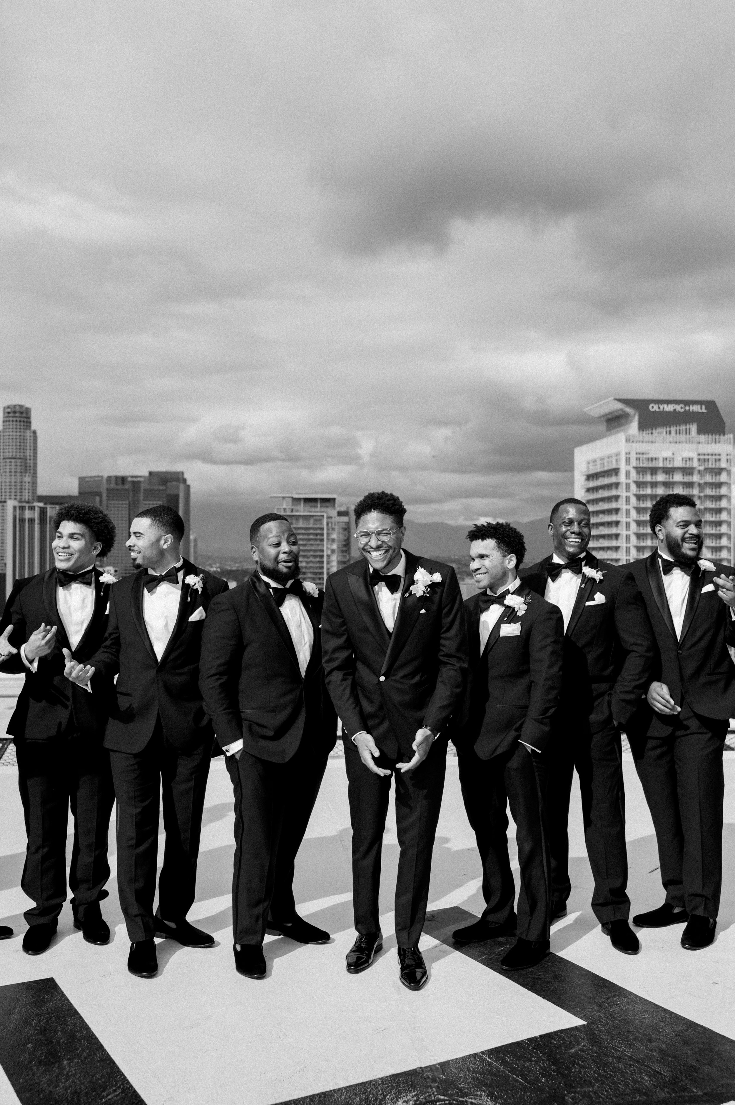 Black and white photo of groomsmen in tuxedos laughing together on rooftop with city skyline in background.