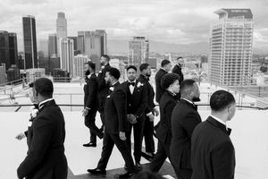 A group of men in formal black suits gather on a rooftop terrace overlooking a city skyline of tall buildings.