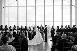 Wedding ceremony taking place in a bright modern venue with large windows and guests seated in black chairs.