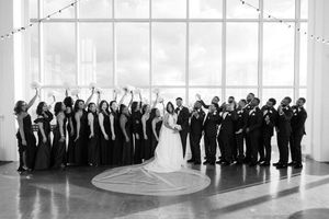 Wedding party celebrates in grand ballroom with dramatic window and decorative string lights overhead in black and white.