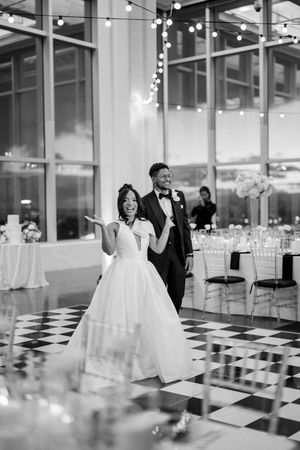 A couple in wedding attire dancing and celebrating in a ballroom with a black and white checkered floor and string lights.