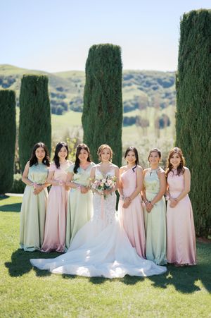 Wedding party in pink and sage green dresses pose together on a grassy lawn with tall cypress trees in the background.