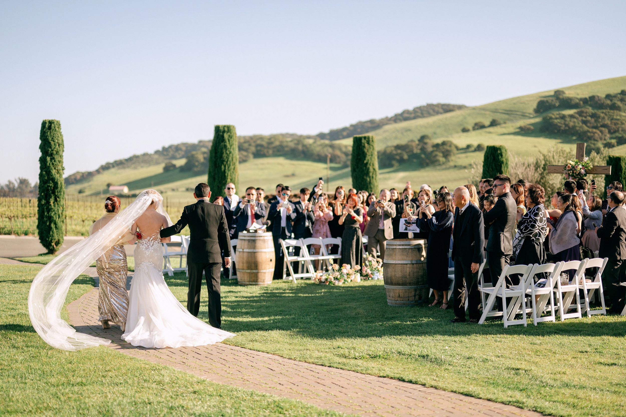 Picturesque outdoor wedding ceremony on a rolling green hillside with white chairs and tall cypress trees.