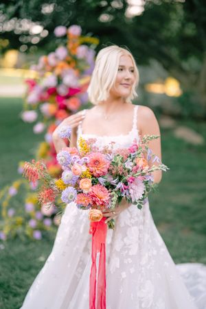 A bride in a white lace gown holds a vibrant bouquet of pink, coral and purple flowers with trailing ribbons in a garden.