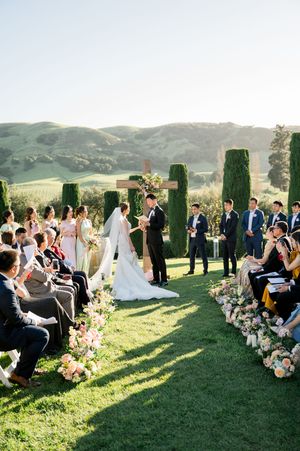 An outdoor wedding ceremony with guests seated on white chairs surrounded by floral arrangements and cypress trees.