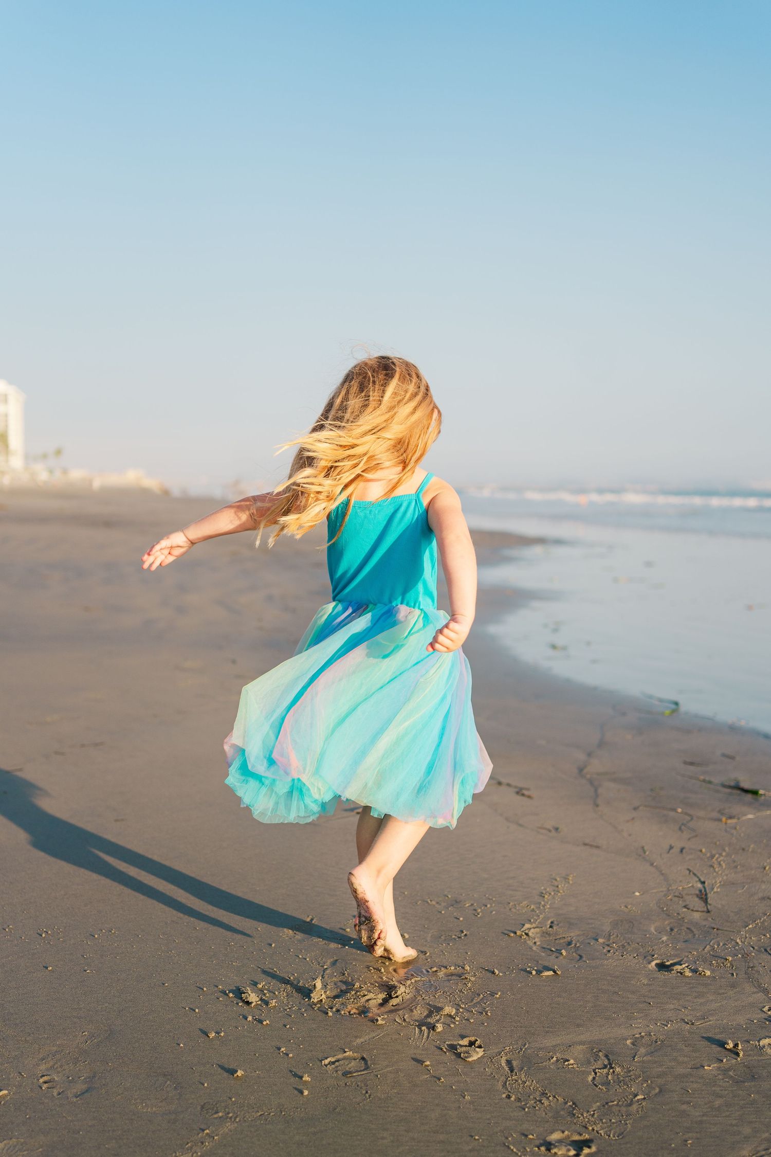 A person in a flowing turquoise dress twirls and dances carefree on a sunny beach by the ocean.