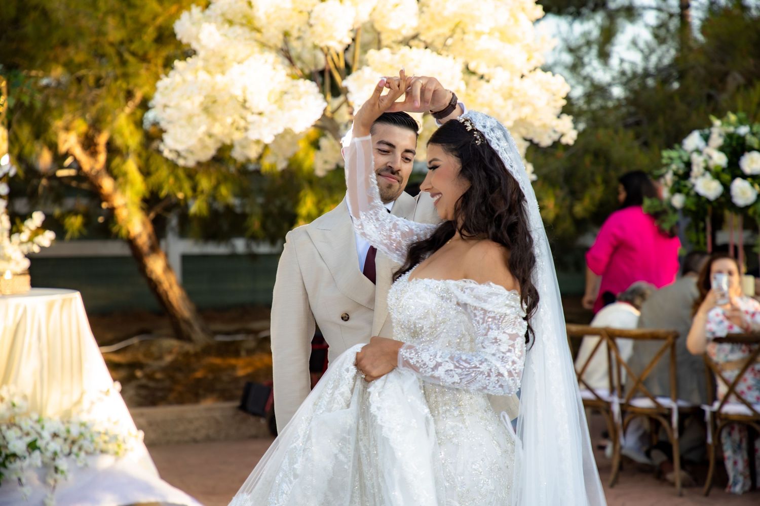 Bride in lace wedding gown and groom in white suit dance under white floral arch outdoors.