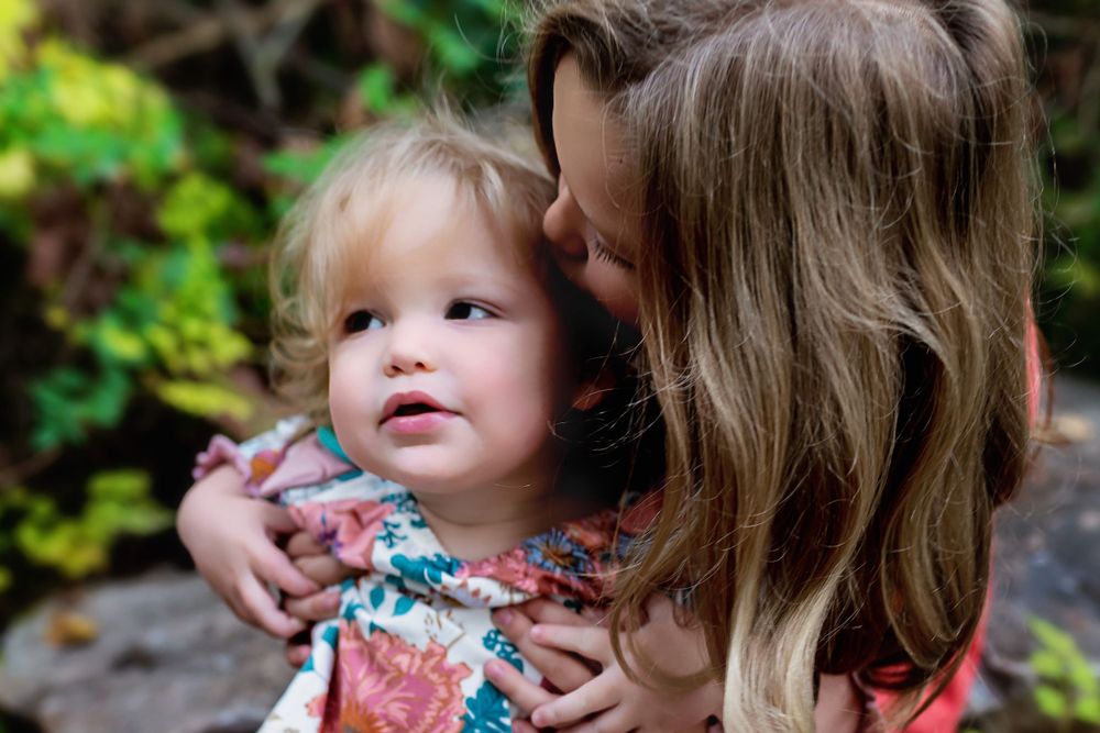 Close-up tender moment between parent and toddler against green foliage.
family photography