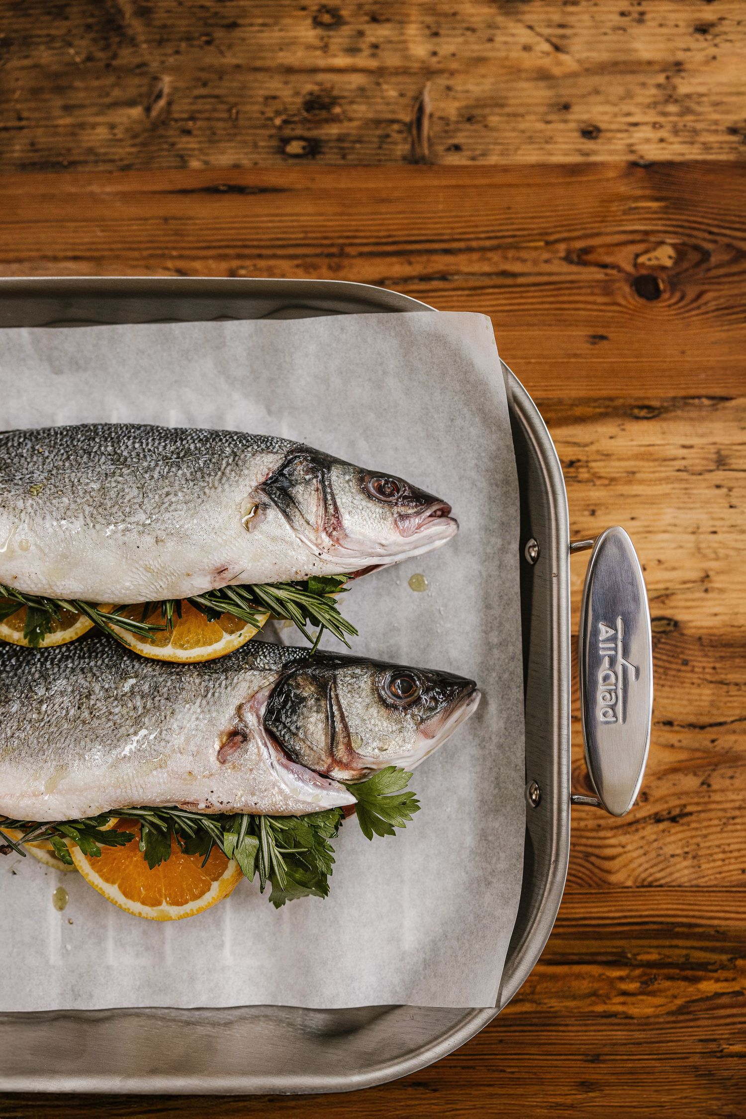 Two fresh whole sea bass fish on parchment paper with lemon slices and herbs in an All-Clad roasting pan.