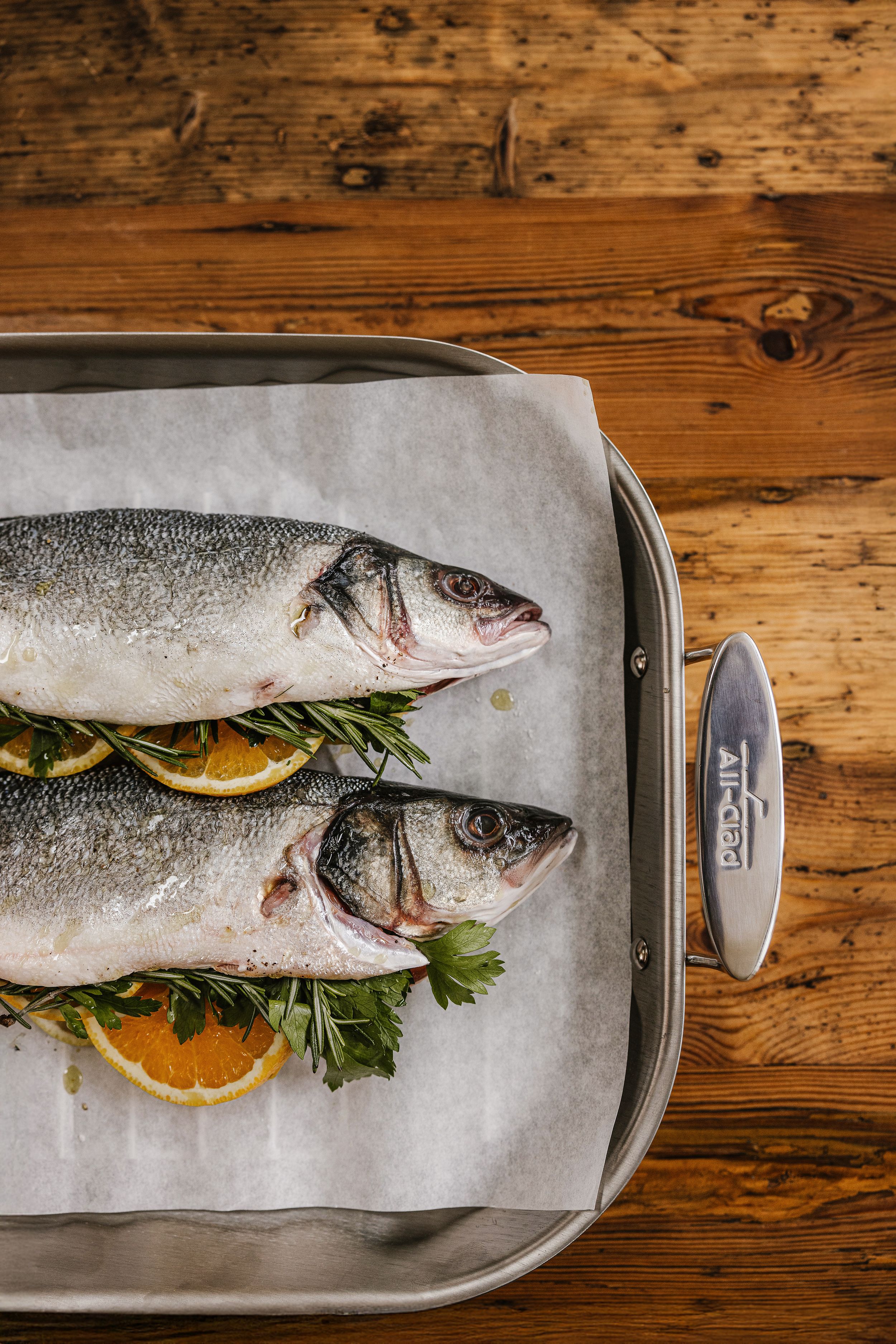 Two fresh whole sea bass fish on parchment paper with lemon slices and herbs in an All-Clad roasting pan.
