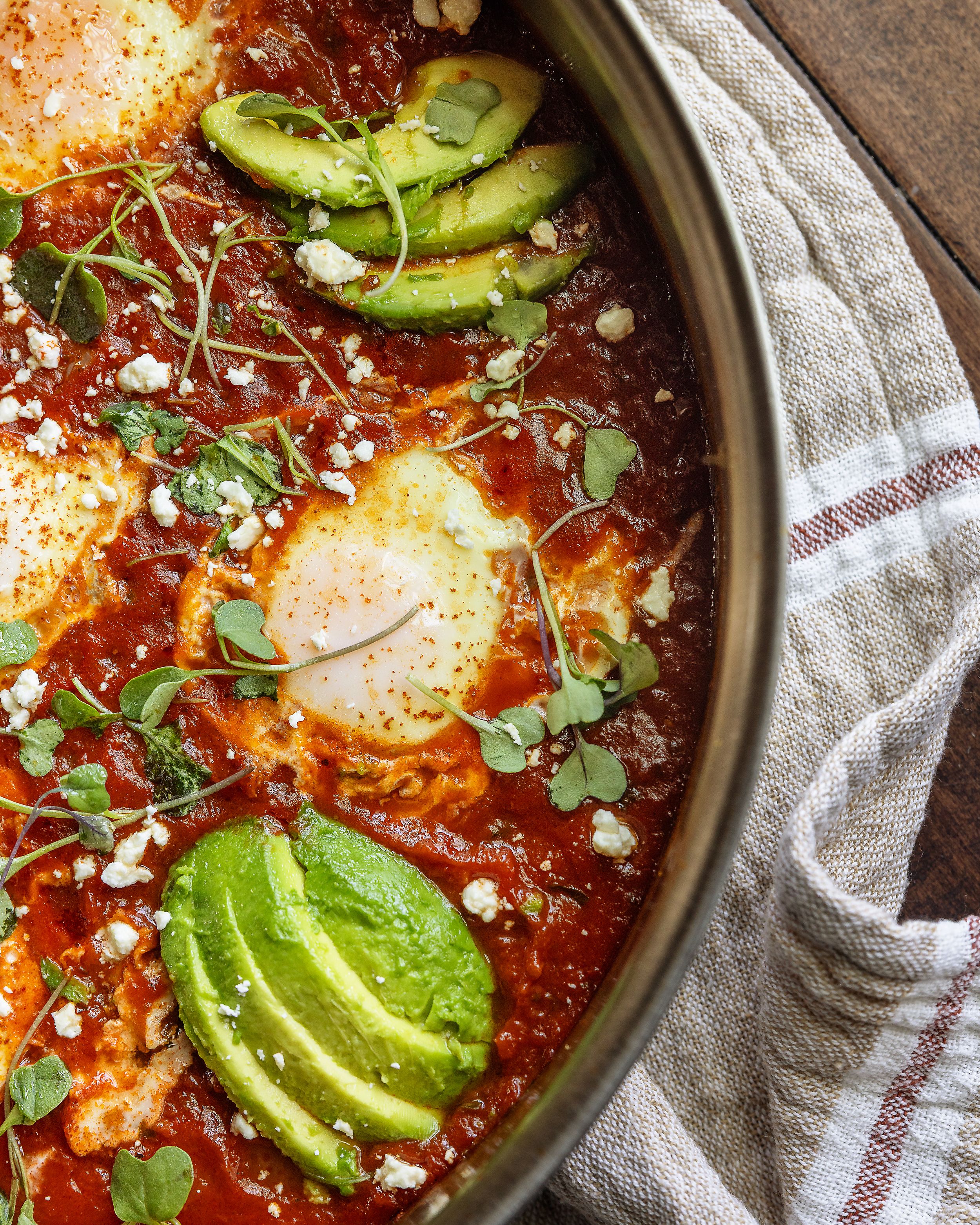 Shakshuka with poached eggs in tomato sauce topped with avocado, feta, and fresh herbs.