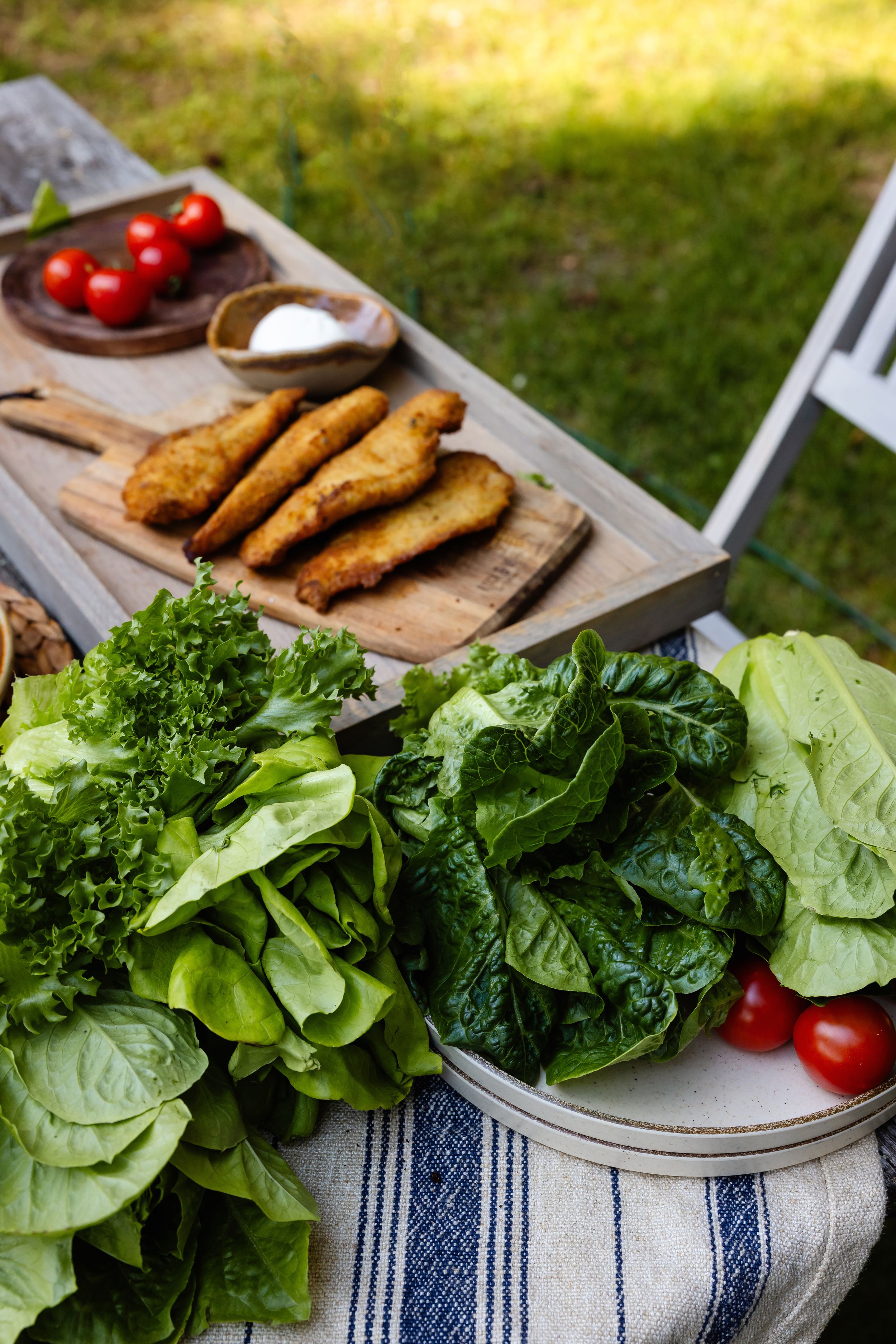 Outdoor table spread with fresh green salad leaves, cherry tomatoes, and golden fried chicken tenders.